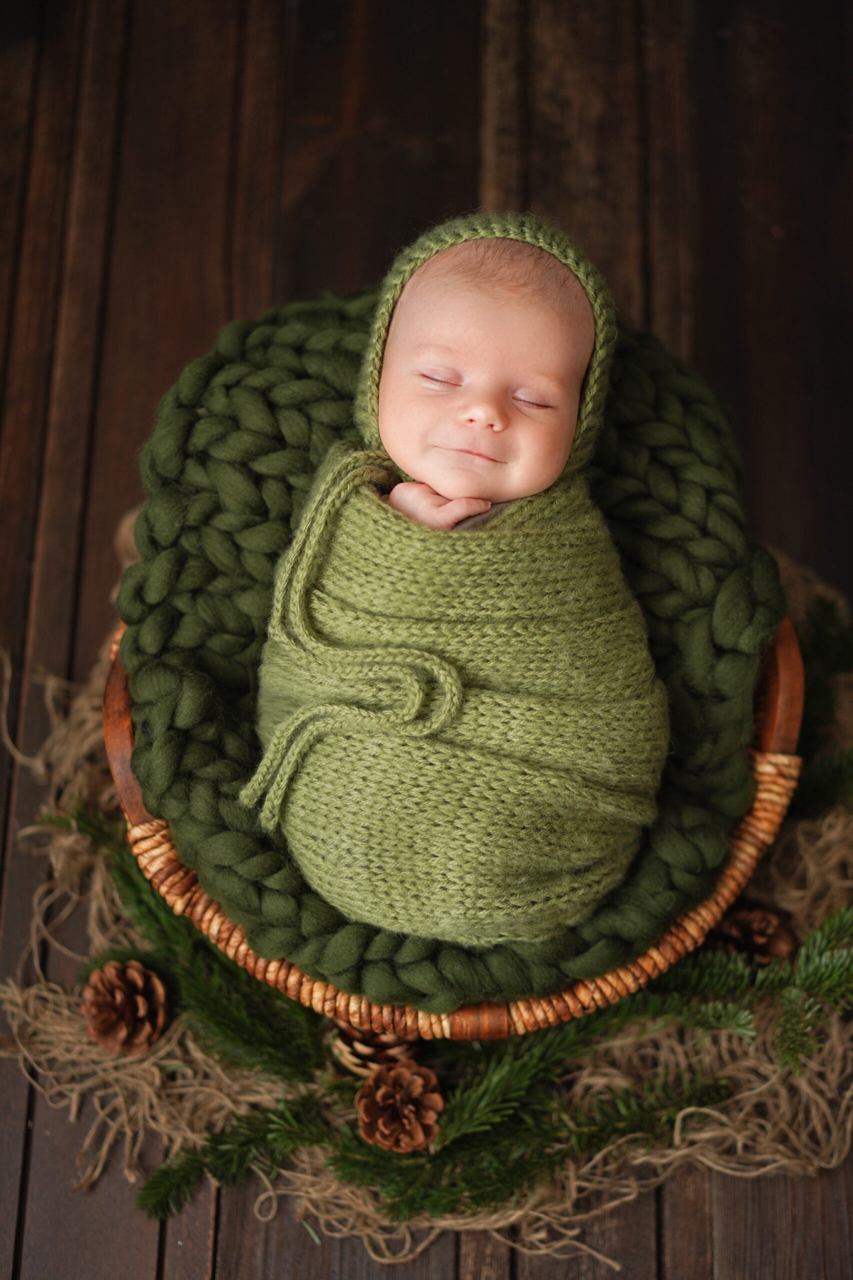 Newborn baby in a dark green swaddle, sleeping in a rustic wooden basket.