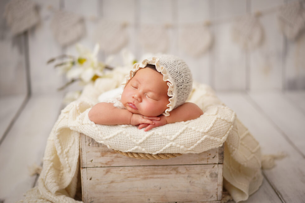 Pasgeboren baby met een wit kanten mutsje, slapend in een crèmekleurige mand met een vachtvoering.